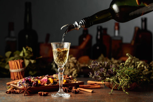 La liqueur ou le mélange à base de plantes jaunes est versé d'une bouteille vintage dans un verre. Sur une table des herbes séchées, des fleurs, des épices et de vieux ustensiles de cuisine. Concept de phytothérapie. - Photo, image