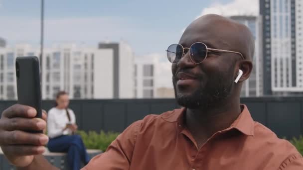 Boczna klatka piersiowa do góry nagranie pozytywnego Afroamerykanina brodatego mężczyzny w okularach przeciwsłonecznych i słuchawkach patrzącego na ekran telefonu i mówiącego podczas rozmowy wideo w nowoczesnym parku na dachu latem - Materiał filmowy, wideo