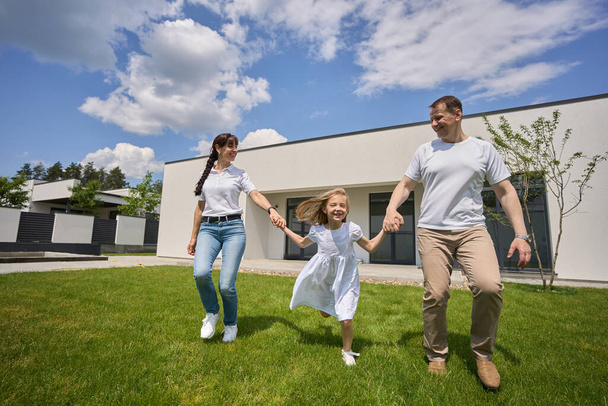 Pleased caucasian mother, father and little daughter holding hands on green yard near modern townhouse in warm sunny summer day. Happy family relationship and spending time together - Photo, Image