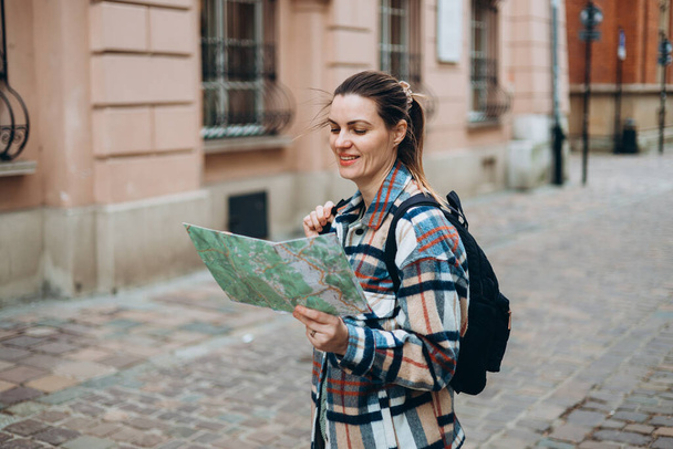 Счастливая жизнерадостная молодая женщина с картой, идущая по городской улице, концепция городского образа жизни. Путешественник. Привлекательная туристка с рюкзаком исследует новый город - Фото, изображение