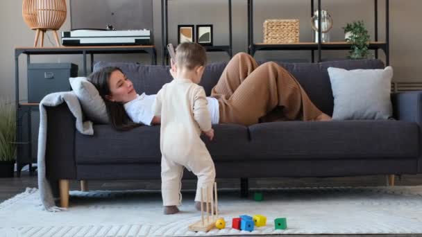 Šťastná mladá maminka leží na pohovce, odpočívá s mobilním telefonem a pozoruje svého batole syn hraje s hračkami a směje se v obývacím pokoji. - Záběry, video