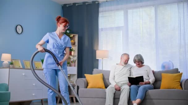 pomoc starszemu, młodemu pracownikowi opieki społecznej w mundurze medycznym z odkurzaczem w rękach pomaga staruszkowi i kobiecie w sprzątaniu mieszkania - Materiał filmowy, wideo