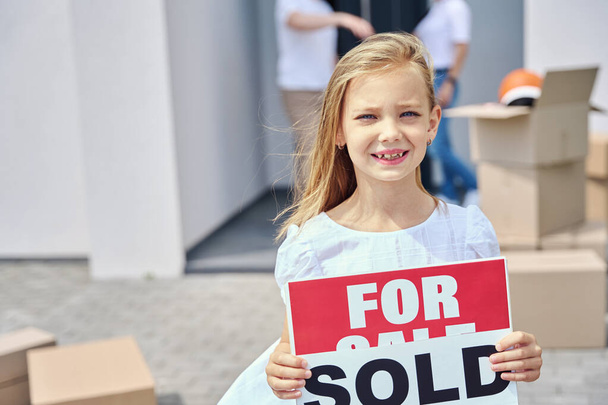 Linda chica se para en el patio con letreros en venta, vendidos, alrededor de una caja con cosas - Foto, Imagen
