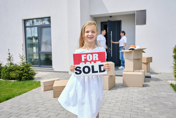 Meisje staat op de binnenplaats van een landhuis met borden te koop, verkocht, haar ouders staan op de veranda - Foto, afbeelding