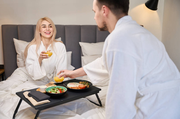 Mladý manžel přinesl podnos se snídaní své ženě v posteli, novomanželé odpočívají v pohodlném pokoji - Fotografie, Obrázek