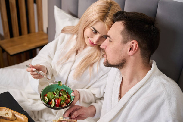 Мила блондинка і її чоловік снідають в комфортабельному готельному номері, вони знаходяться в затишній спальні - Фото, зображення