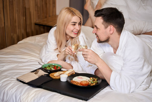 Pasgetrouwden genieten van ontbijt op bed met champagne, ze zijn in donzige badjassen - Foto, afbeelding