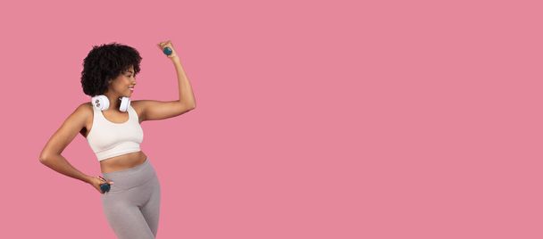 Itsevarma nuori musta nainen kunto harrastaja taivuttaa hauiksensa pitäen käsipaino, kuulokkeet kaulassaan vastaan elinvoimainen vaaleanpunainen tausta, panoraama vapaata tilaa - Valokuva, kuva