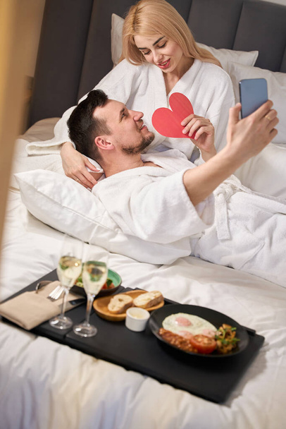 Paar macht ein Selfie in der Flitterwochen-Suite mit süßem Geschenkherz, sie frühstücken mit Champagner - Foto, Bild