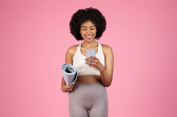 Улыбающаяся, стройная молодая черная женщина в спортивной одежде держит коврик для йоги, счастливо переписываясь на смартфоне, стоя на розовом фоне студии - Фото, изображение