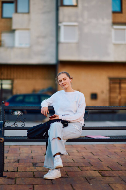 Uma linda adolescente loira se senta em um banco de parque, usando seu smartphone e fones de ouvido sem fio para se preparar para a escola, com sua mochila escolar ao lado dela no banco.  - Foto, Imagem