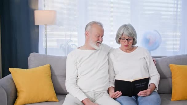 caring for pensioners, an elderly married couple enjoys relaxing and reading book while sitting on sofa while a female social worker helps them with cleaning room - Footage, Video