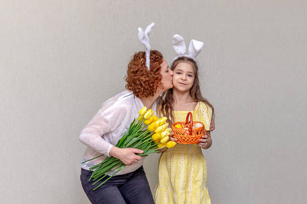 A woman with flowers and a girl with bunny ears and a basket of Easter eggs. The family celebrates Easter - Photo, Image