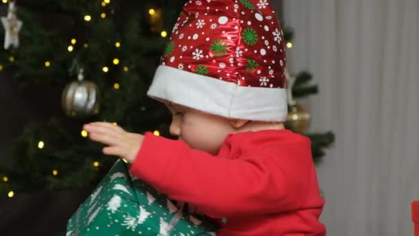 Jeden-rok-starý kluk v Santa klobouk otevírá dárky u vánočního stromečku. - Záběry, video