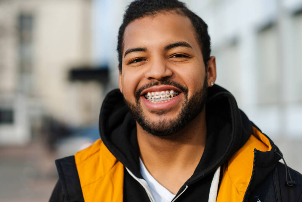 Gülümseyen, yakışıklı, diş teli takan, ceketli, şehir sokağında poz veren ve kameraya bakan Afro-Amerikan bir öğrencinin portresi. - Fotoğraf, Görsel
