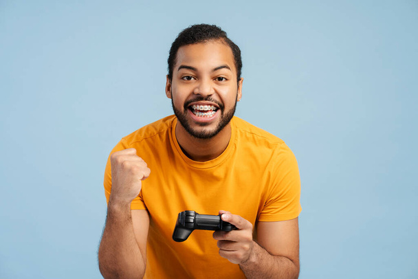 Portrait of a joyful African American man with braces, playing a video game and holding a joystick, making a victory gesture with fist and looking at the camera, isolated on a blue background - Photo, Image