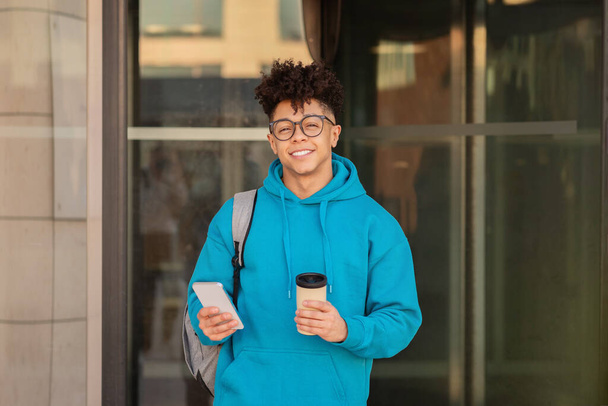 Молодой чёрный мужчина в очках делает перерыв на кофе, неторопливо просматривая свой смартфон в городской обстановке, улыбаясь в камеру. Счастливый студент, стоящий у здания колледжа - Фото, изображение