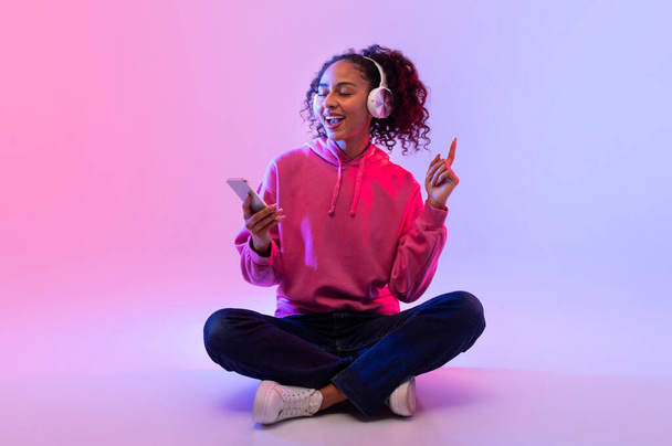 Smiling black lady with curly hair, sitting cross-legged, enjoys music on headphones, feeling the rhythm with finger in the air while holding smartphone against pink gradient background - Photo, Image