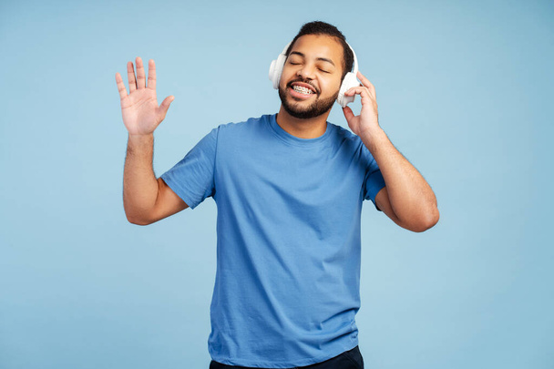 Wesoły, atrakcyjny Afroamerykanin z aparatem na zęby, noszący słuchawki, tańczący i cieszący się muzyką z zamkniętymi oczami, odizolowany na niebieskim tle - Zdjęcie, obraz