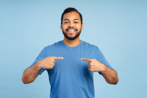 Gülümseyen Afro-Amerikan bir erkek öğrencinin resmi, günlük kıyafetlerle, diş telleriyle, gururla kendini gösteriyor, kameraya bakıyor, mavi bir arka planda izole edilmiş. - Fotoğraf, Görsel