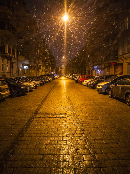 Wroclaw, Poland - November 25 2023: Beautiful and long street with brick and lanterns hanging over the center of street and cars parked on both sides at snowy night - Photo, Image