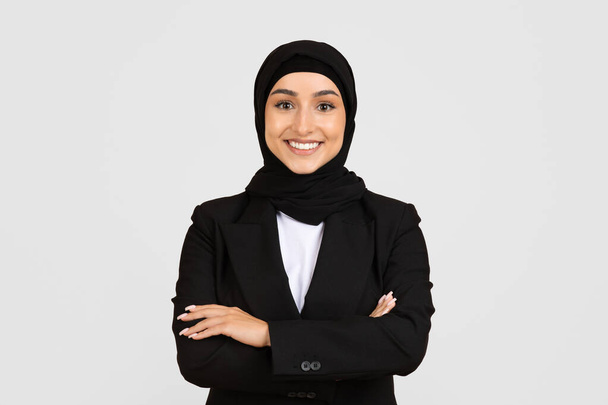 Jeune femme souriante confiante et professionnelle portant hijab et costume d'affaires noir avec les bras croisés debout sur fond clair et souriant à la caméra - Photo, image