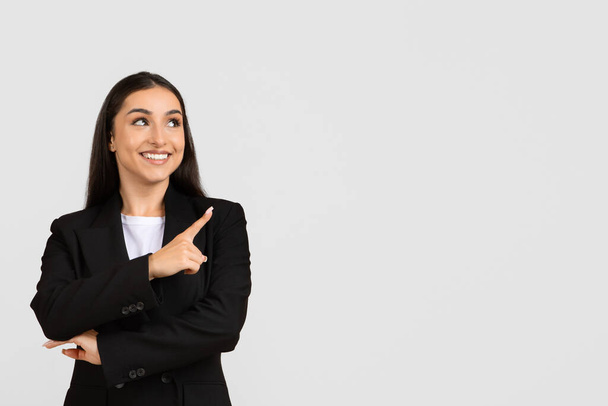 Cheerful young businesswoman in black suit pointing upwards at free space, indicating new idea or business concept on grey background, banner - Photo, Image