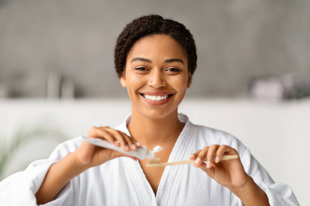 Vidám fekete nő alkalmazó fogkrém bambusz fogkefe, mosolygós afro-amerikai nő fürdőköpenyben készül a szájhigiénia otthon, áll a modern fürdőszoba belső - Fotó, kép