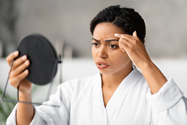 Besorgte schwarze Frau schaut im Handspiegel auf Pickel auf der Stirn, besorgte afrikanisch-amerikanische Frau trägt Bademantel fleckende Haut Problem, während Beauty-Routine im modernen Badezimmer - Foto, Bild