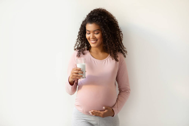 Güzel Afrikalı Amerikalı anne elinde bir bardak süt ve gülümseyen mutlu siyah hamile kadın besin sağlığına ve bebeğinin refahına odaklanıyor, beyaz arka planda duruyor. - Fotoğraf, Görsel