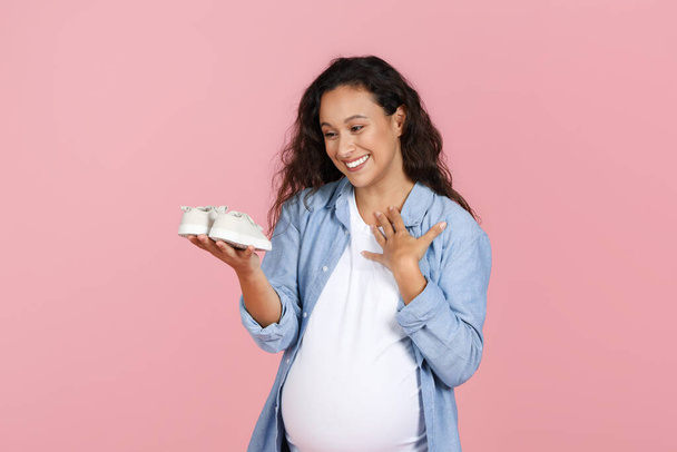 Moederschap concept. Opgewonden zwangere vrouw met grote buik kijken naar baby schoenen op haar handpalm en aanraken van haar borst, emotioneel gevoel, verwacht kind, roze studio achtergrond - Foto, afbeelding