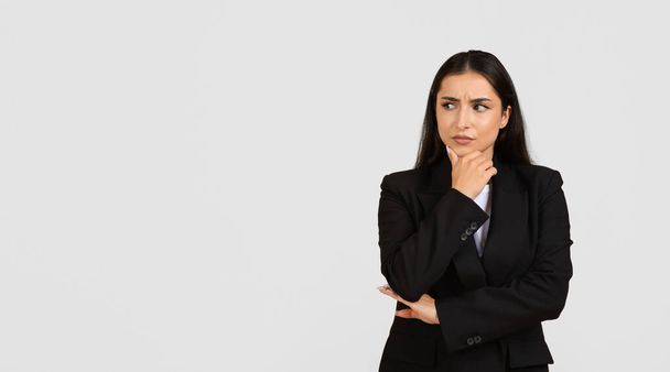 Jeune femme professionnelle contemplative en costume d'affaires noir regardant de côté l'espace libre et touchant le menton, sur fond gris, panorama, bannière - Photo, image
