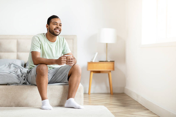 Χαρούμενη ελκυστική χιλιετή αφροαμερικανός άνδρας κάθεται στο κρεβάτι στο σπίτι, πίνοντας καφέ, κοιτάζοντας μέσα από το παράθυρο και χαμογελώντας, απολαμβάνοντας ηλιόλουστο πρωινό, αντίγραφο χώρο για διαφήμιση - Φωτογραφία, εικόνα