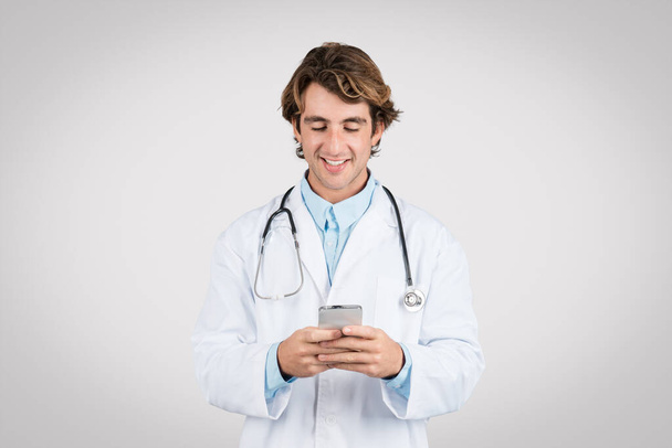 Улыбающийся молодой врач-мужчина со стетоскопом вокруг шеи счастливо печатает на смартфоне, возможно, общаясь с пациентами или коллегами онлайн - Фото, изображение