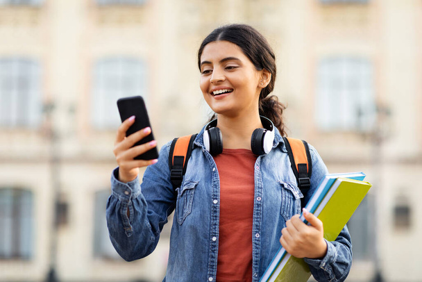 Çevrimiçi öğrenme, e-eğitim konsepti. Açık havada telefon kullanan, sırt çantası ve ders kitapları taşıyan mutlu Hintli genç kız öğrenci üniversiteye gidiyor, internetten programını kontrol ediyor. - Fotoğraf, Görsel