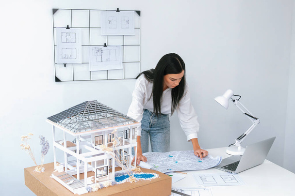 Portrait of Young beautiful caucasian architect focus on comparing house structure between house model and blueprint and laptop placed on meeting table. Check, inspect, estimate concept. Immaculate. - Photo, Image