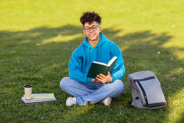 Навчальний пікнік. Веселий чорний студент в окулярах читає книгу, сидить на зеленій траві, вчиться з кавою на відкритому повітрі в парку, посміхаючись камері. Навчання на газоні у вихідні - Фото, зображення