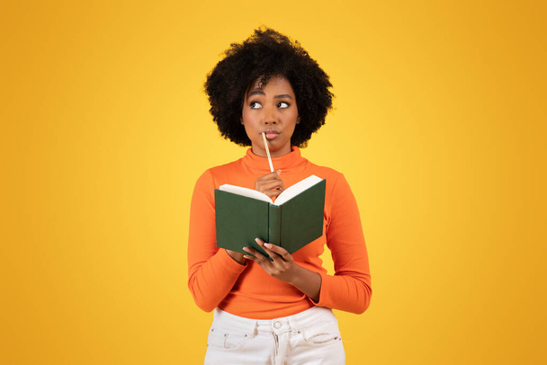 Nachdenkliche junge schwarze Frau mit lockigem Haar, die beim Lesen eines grünen Buches nachdenklich einen Bleistift ans Kinn hält, gekleidet in ein orangefarbenes Oberteil mit gelbem Hintergrund, Studio - Foto, Bild