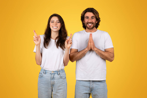Hopeful european young woman with crossed fingers and cheerful man with palms together in a gesture of wishful thinking, both in white t-shirts against a yellow background - Photo, Image