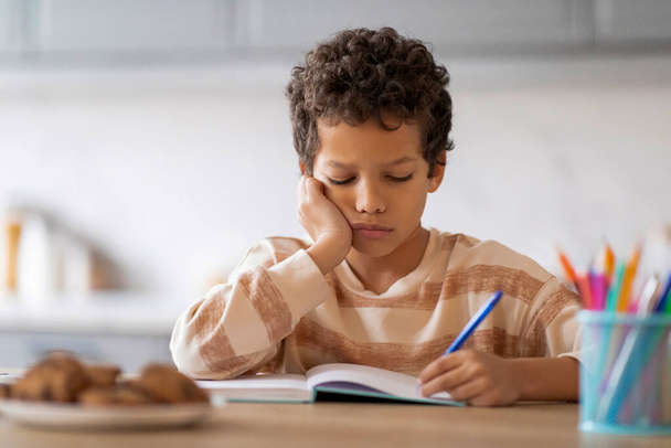 Little black boy feeling tired and sleepy while doing his homework, exhausted african american preteen schoolboy sitting at desk in kitchen and falling asleep while writing in notebook, copy space - Fotó, kép