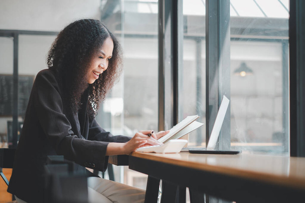Сфокусированная деловая женщина с вьющимися волосами занимается своей работой в современном кафе, используя ноутбук и делая заметки в спокойной, хорошо освещенной обстановке.. - Фото, изображение