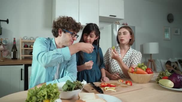 Attractive caucasian father and mother spend time together to cook fresh meal with asian daughter. Dad and mom preparing fresh vegetable for making salad at kitchen and eating vegetable. Pedagogy. - Footage, Video