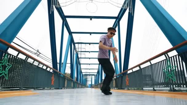 Hipster provádět b-boy kroky a street dance na mostě s pozadím. Profesionální break dance nosit stylové oblečení a dívat se na kameru, zatímco pózovat na kameru. Venkovní sport2024. Vesele. - Záběry, video