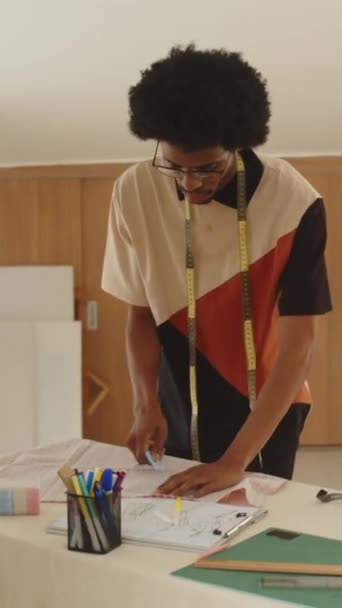 Vertikale Aufnahme eines afroamerikanischen männlichen Schneiders, der ein Stück Stoff markiert, bevor er das Kleid im Atelier näht - Filmmaterial, Video