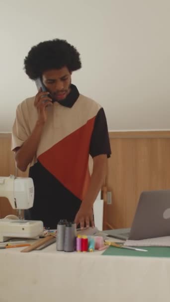Tiro vertical de costurero negro llamando al cliente en el teléfono inteligente y utilizando el ordenador portátil en el estudio de costura - Metraje, vídeo