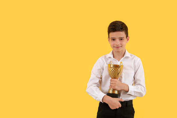 イエローバックグラウンドで賞金カップを持つスマートな小さな学校の少年 - 写真・画像