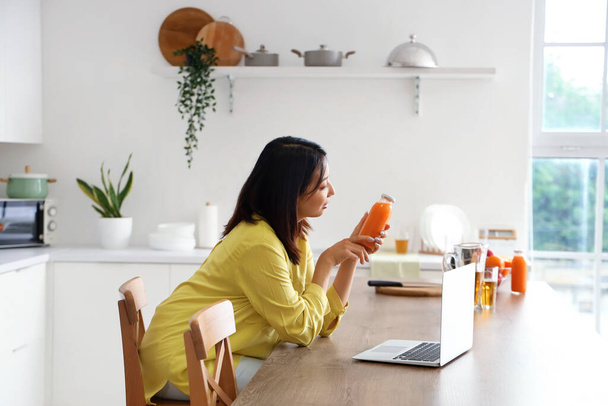 Beautiful Asian woman with bottle of juice and laptop in kitchen - Φωτογραφία, εικόνα