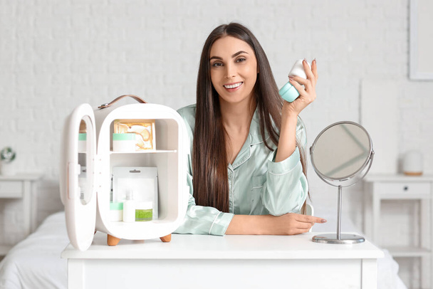 Schöne junge glückliche Frau mit Gläschen Sahne und kleinem Kühlschrank für Kosmetikprodukte auf dem Tisch im Schlafzimmer sitzend - Foto, Bild
