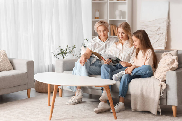 Niña con su mamá y su abuela leyendo la revista en el sofá en casa - Foto, Imagen