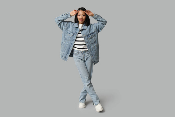 Красива молода щаслива азіатка в стильному джинсовому одязі на сірому фоні - Фото, зображення